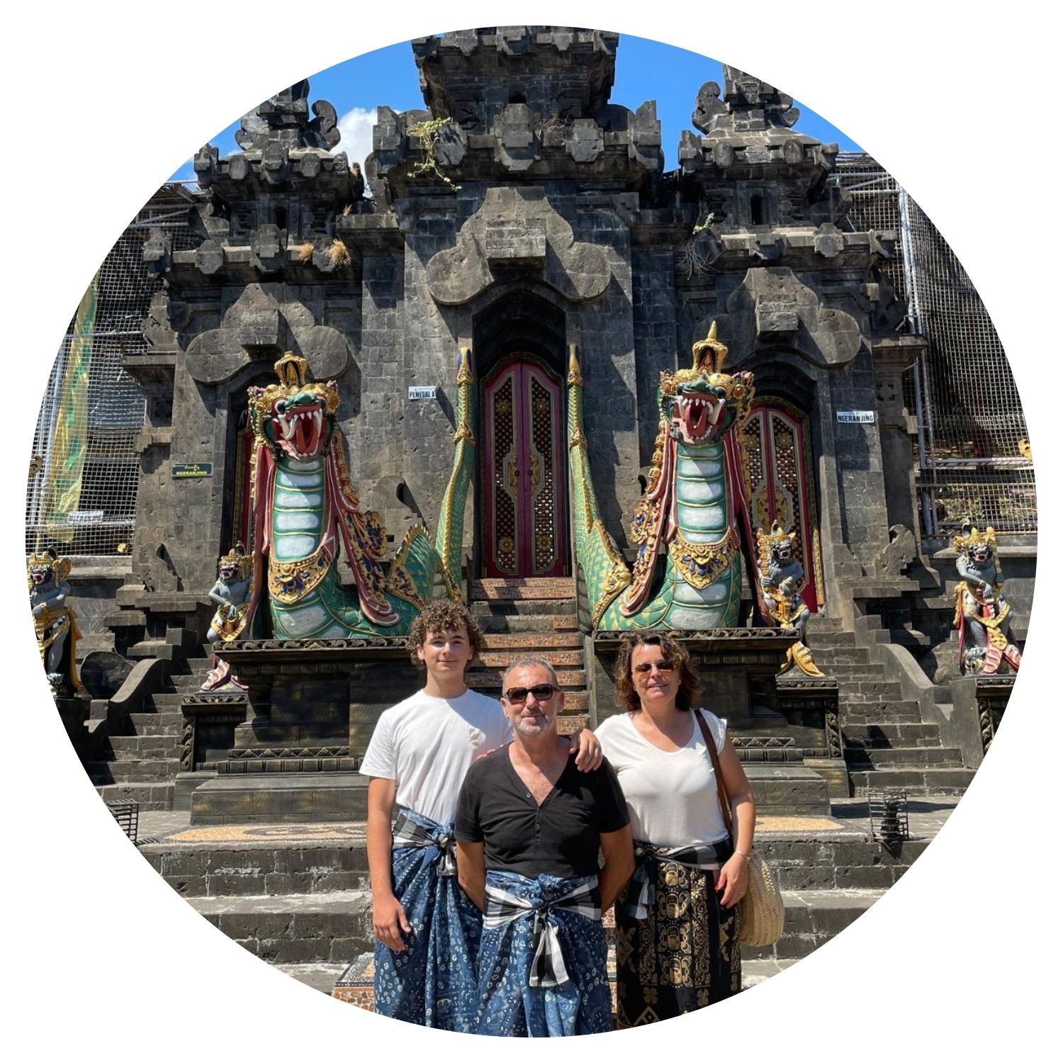 Une famille de trois personnes, deux adultes et un adolescent, pose devant l'entrée ornée d'un temple à Bali. Ils portent des sarongs traditionnels, signe de respect dans les lieux sacrés balinais. Derrière eux, l'architecture du temple est richement décorée avec des sculptures colorées de dragons.