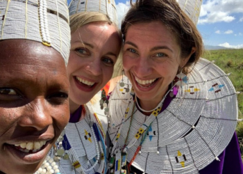 Trois femmes souriantes portant des colliers traditionnels Massaï en Tanzanie, capturées sous un ciel ensoleillé.