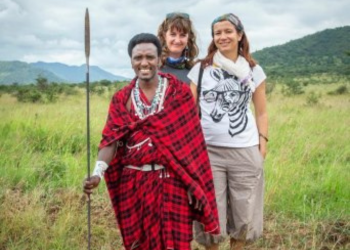 Guerrier masaï en tenue traditionnelle rouge posant avec deux femmes occidentales dans la savane tanzanienne.