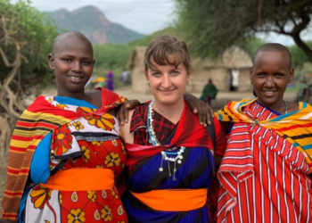 Trois femmes souriantes - deux Tanzaniennes en tenues traditionnelles colorées et une femme occidentale au centre - posant ensemble dans un village tanzanien.