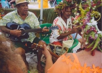 Groupe de musiciens polynésiens en train de jouer de la musique traditionnelle, portant des chemises colorées et des couronnes de fleurs.