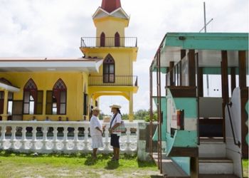 Deux personnes se tiennent devant une église colorée en Polynésie, discutant paisiblement. À droite, un bâtiment en bois ajoute une touche traditionnelle à la scène.