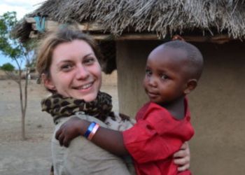 Femme souriante tenant un enfant tanzanien devant une habitation traditionnelle.