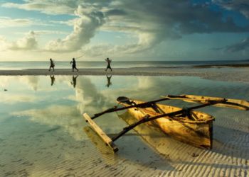 Un bateau traditionnel en bois échoué sur une plage de Zanzibar avec trois personnes marchant au loin.